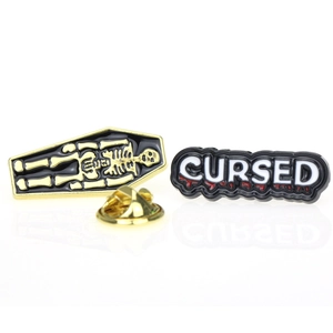 Macro shot of coffin-shaped skull pin and 'CURSED' text pin with red enamel drip