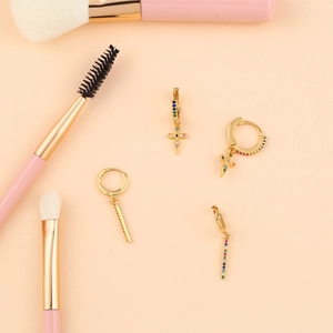 Detail of gold cross and bar earrings with colorful zircons next to makeup brushes