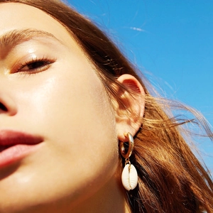 Close-up of a shell charm earring on a gold hoop worn against a bright blue sky background
