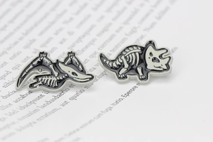 Macro detail of dinosaur skeleton enamel pins on book pages showing pterosaur and triceratops