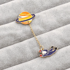 Detail of cartoon astronaut and Saturn enamel pins connected by a fine gold metal chain