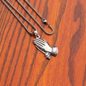 Close-up of silver stainless steel praying hands pendant on a box chain over wood background