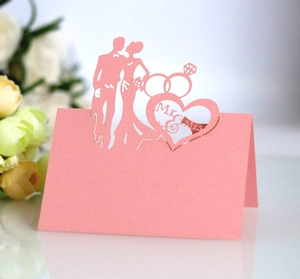 Close-up of pink laser-cut wedding place card with Mr & Mrs silhouette and heart ring design