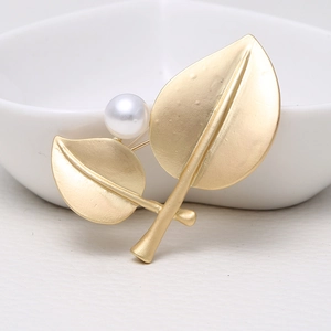 Elegant gold leaf-shaped brooch with a white pearl accent on a minimalist ceramic surface