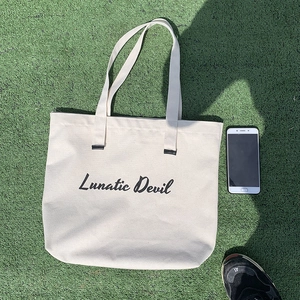 Flat lay detail of a beige canvas tote bag with Lunatic Devil text on green grass
