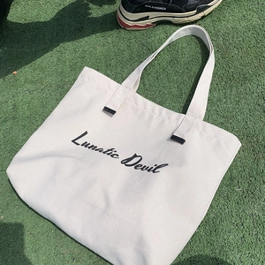 Detail of off-white canvas tote bag with Lunatic Devil script on a green grass background
