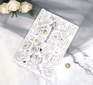 Close-up of white laser-cut floral invitation cover on marble with white rose and jewelry