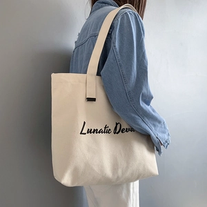 Detail of a beige canvas tote bag with black Lunatic Devu lettering shown on shoulder