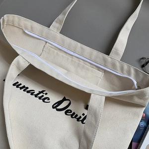 Beige canvas tote bag with Lunatic Devil print showing top zipper and inner pocket detail