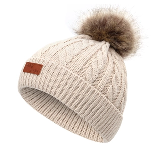 Détail d'un bonnet en maille torsadée beige avec pompon en fausse fourrure et étiquette cuir