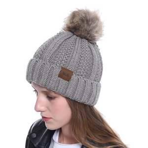 Detail of grey chunky knit beanie featuring a brown faux fur pompom and leather brand tag