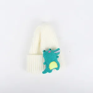 Detailansicht der weißen Kinderstrickmütze mit einem niedlichen grünen Dino-Plüschaufnäher