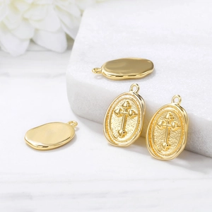 Gold oval metal charms with embossed cross and heart designs on marble surface