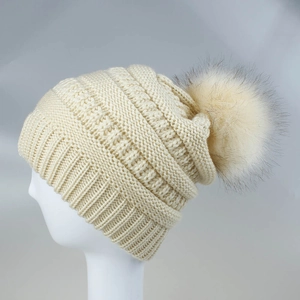 Detail einer beigen Strickmütze mit Zopfmuster und einem großen flauschigen Kunstfellbommel