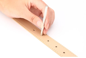 Close-up of a hand using a metal punch tool on a beige leather belt with fine stitching
