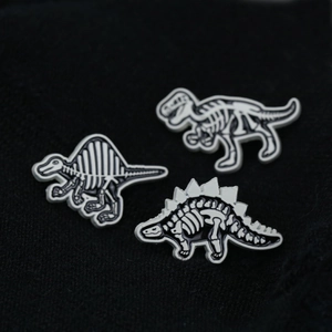 Three dinosaur skeleton enamel pins in black and white finish on dark fabric