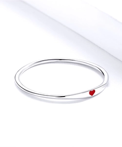 Dettaglio di un bracciale rigido in argento con un piccolo cuore rosso su metallo liscio