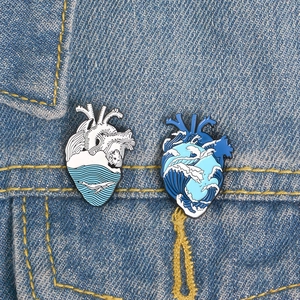 Anatomical heart enamel pins with whale and ocean wave designs on denim fabric