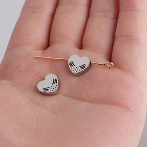 Polished stainless steel hollow heart beads with angel wings shown on a needle in hand