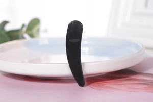 Detail of a curved black facial cosmetic spatula resting on a ceramic dish for cream