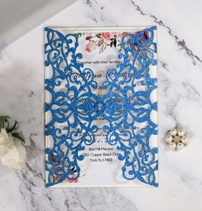 Detailed laser-cut blue glitter wedding invitation on white marble with pearl accents