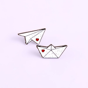 Detail of white enamel paper plane and boat brooches with red heart on purple background