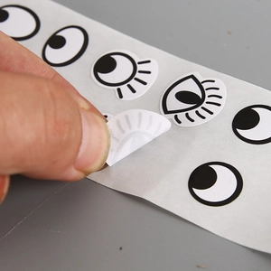 Roll of colorful cartoon eye stickers for DIY crafts, showing hand peeling adhesive label