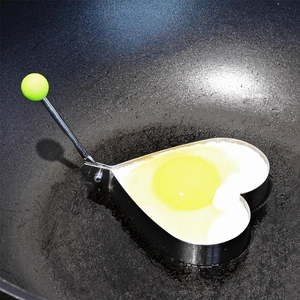Heart-shaped stainless steel egg mold with green handle frying egg in a pan