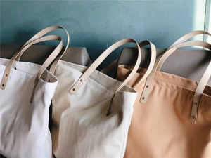 Three minimalist canvas tote bags in white, beige, and tan with leather handles and rivets