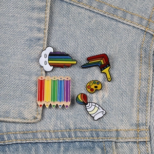 Detail of five rainbow enamel pins on denim jacket showing artistic paint palette and brushes