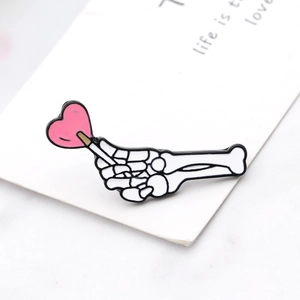 Cartoon enamel brooch featuring a white skull hand making a heart with a pink lollipop