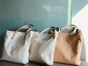 Three minimalist canvas tote bags in white and beige with faux leather handles on a bench