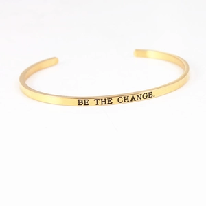 Close-up of gold stainless steel C-shaped bracelet with BE THE CHANGE engraving