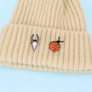 Close-up of enamel snail and ram skull pins pinned on a beige knitted beanie hat