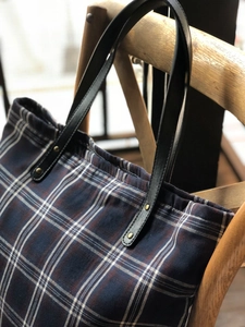 Detail of navy and brick red plaid tote bag with leather handles on a wooden chair