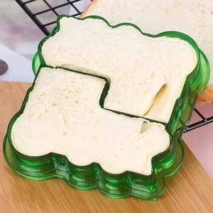 Green house-shaped sandwich cutter mold pressing a white toast slice on a wooden board