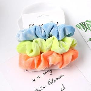 Three-pack velvet hair scrunchies in blue green and orange on a minimalist display