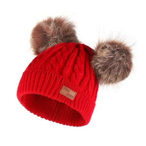 Detail of red cable-knit beanie featuring dual brown fur pom-poms and leather logo patch