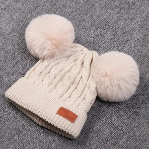 Close-up of beige knit beanie with faux fur double pom-poms and leather logo tag