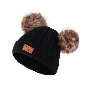 Detail of black knit winter hat featuring twin brown faux fur pom-poms and leather patch