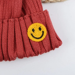Detail of brick red ribbed knit beanie featuring a yellow embroidered smiley face patch
