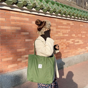 Close-up of a green canvas tote bag with a white patch label against a red brick wall
