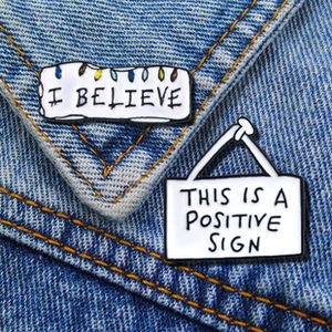 Close-up of I BELIEVE and POSITIVE SIGN enamel pins on blue denim fabric texture