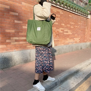 Close-up of a green corduroy tote bag with white label against a vintage brick wall