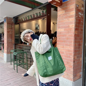 Green canvas tote bag worn by a model in front of a coffee shop, showing capacity and style