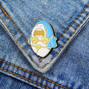 Detail of a cartoon shark enamel pin with gold shell bikini on blue denim fabric