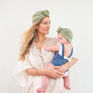 Matching sage green donut turban hats worn by mother and baby against a white wall