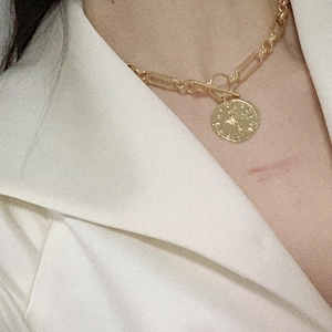 Detalle de collar dorado con colgante de moneda y cierre OT sobre una chaqueta blanca