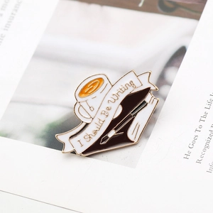 Close-up of enamel pin with coffee cup and book featuring I Should Be Writing text