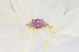 Close-up of golden ring with oval pink gemstone and white zircon on white silk fabric